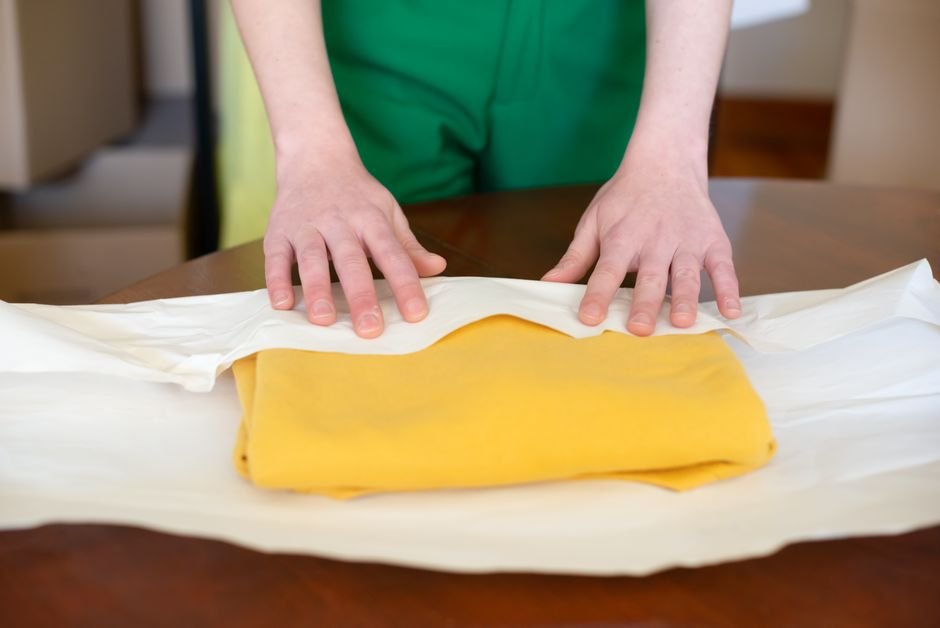 Hands carefully wrapping a yellow garment in paper indoors, emphasizing careful packing.